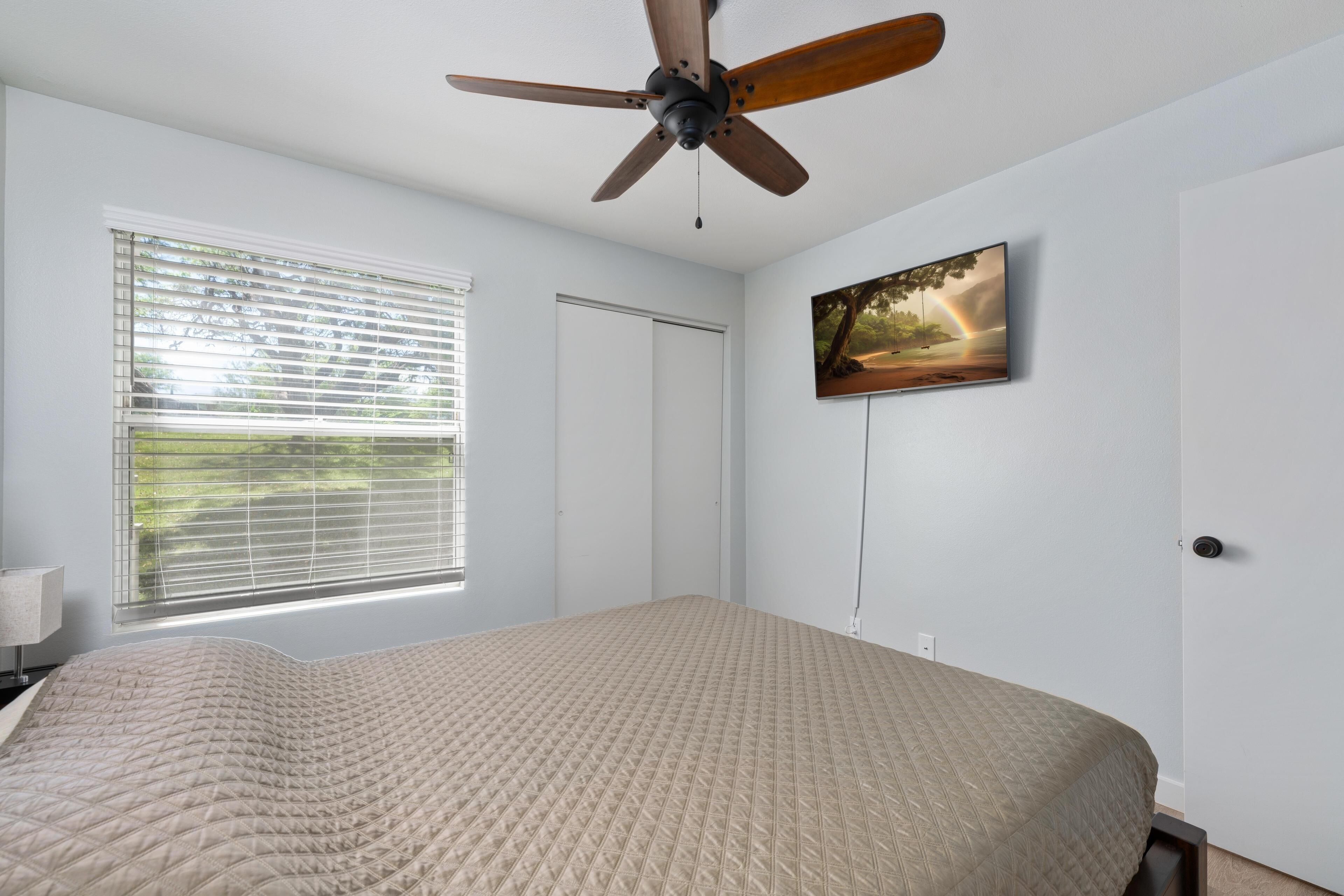A bedroom with a bed, a ceiling fan, a window with blinds, a door, and a painting on the wall.