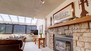 A cozy living room with a stone fireplace, a large window with a view of the mountains, and a comfortable leather sofa. - Vs741 Village Sqaure 3 Bedroom Condo by RedAwning (Copper Mountain)