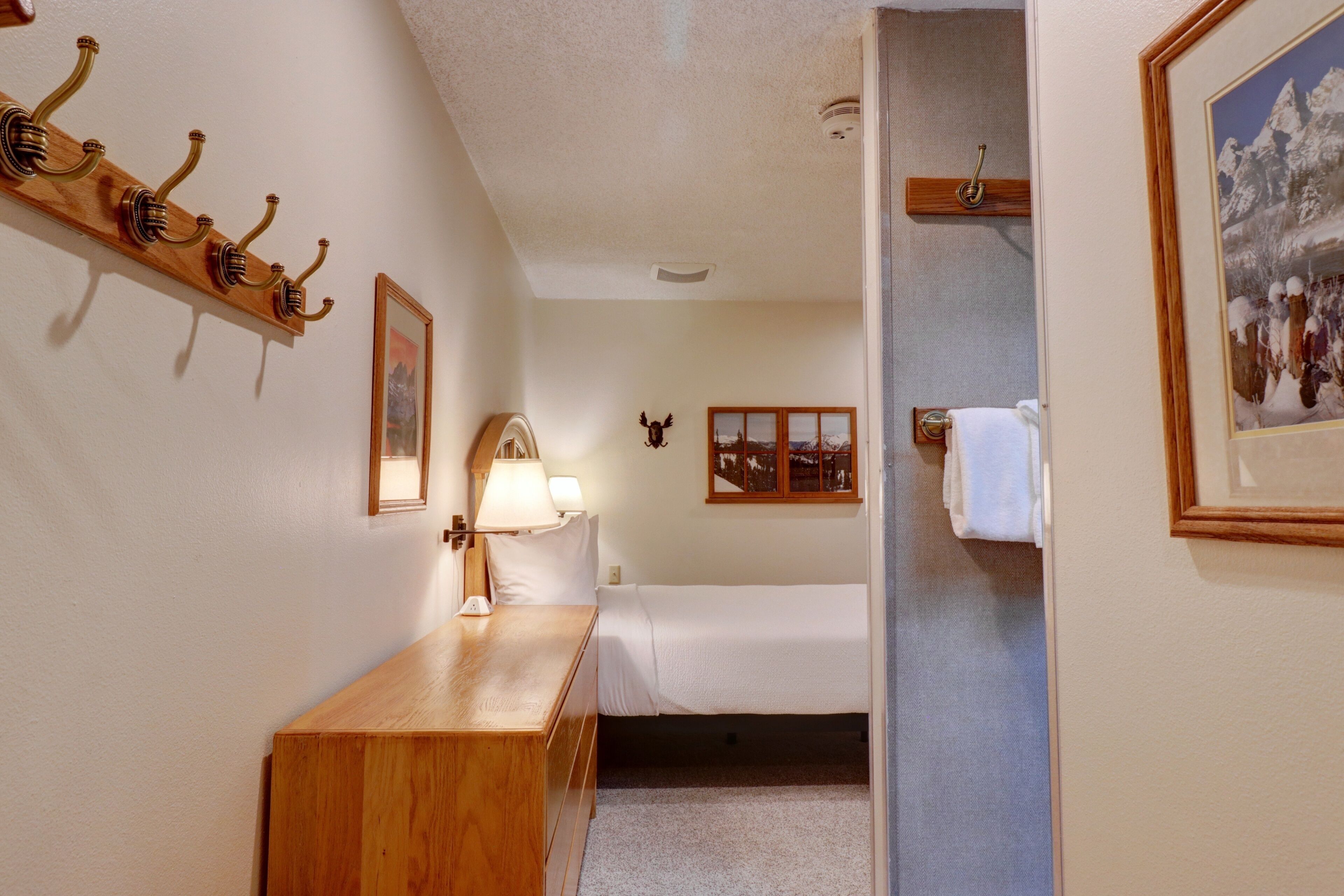 A hotel room with a wooden dresser, a bed, a wall-mounted lamp, a coat rack, and a framed picture of a snowy landscape.