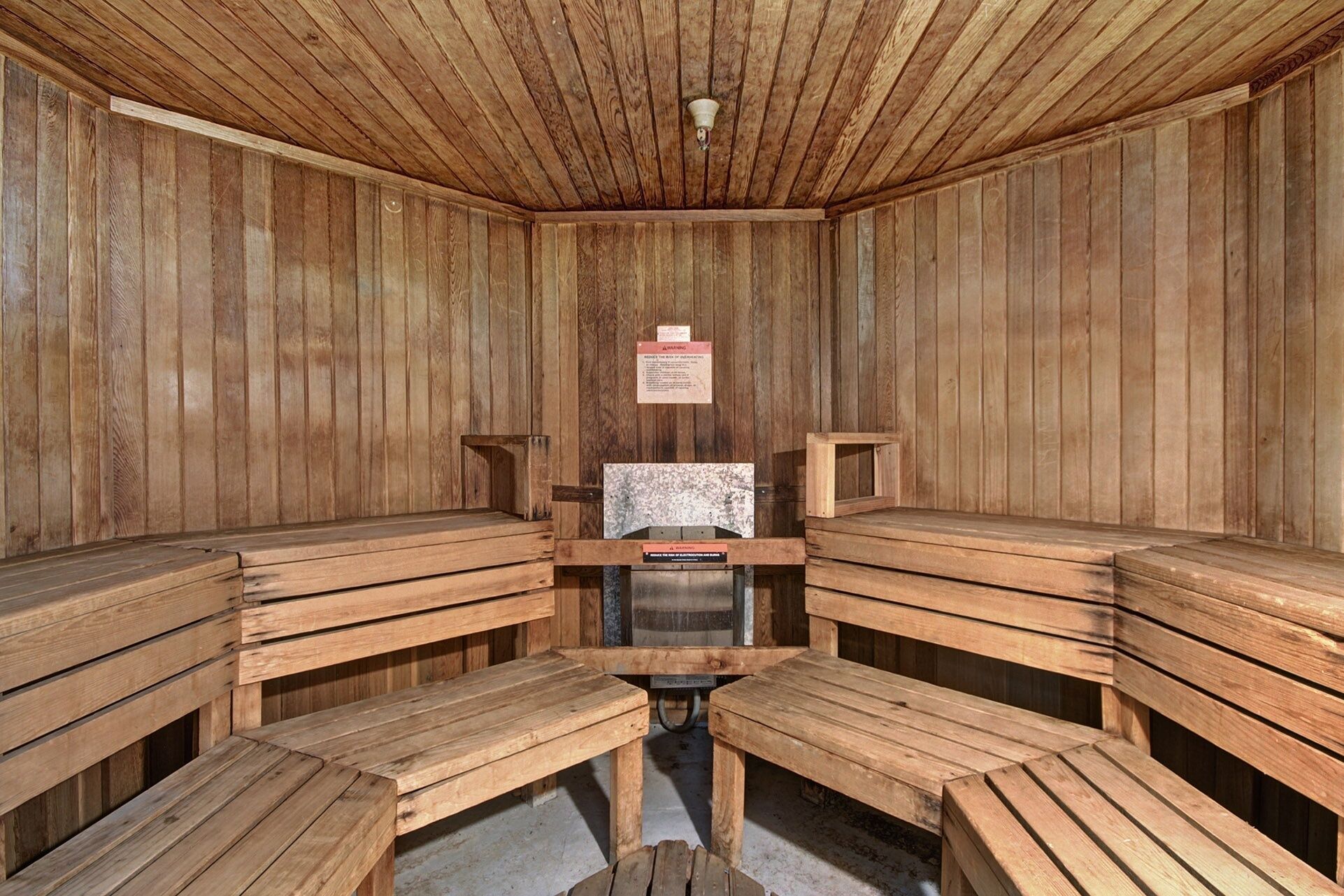 A sauna with wooden benches arranged in a semi-circle, a central heater, and a warning sign on the wall.