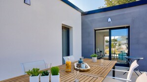A wooden table with potted plants, a glass, and a bottle of orange juice in an outdoor patio area with a modern building in the background. - Modern house with garden - 5min from the beach (Erquy)