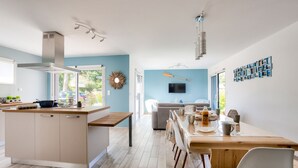 A modern kitchen with a central island, a dining area with a wooden table and chairs, and a living area with a sofa and TV. - Modern house with garden - 5min from the beach (Erquy)
