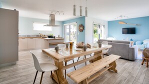 A modern kitchen with a wooden table and benches, a grey sofa, and a flat-screen TV mounted on the wall. - Modern house with garden - 5min from the beach (Erquy)