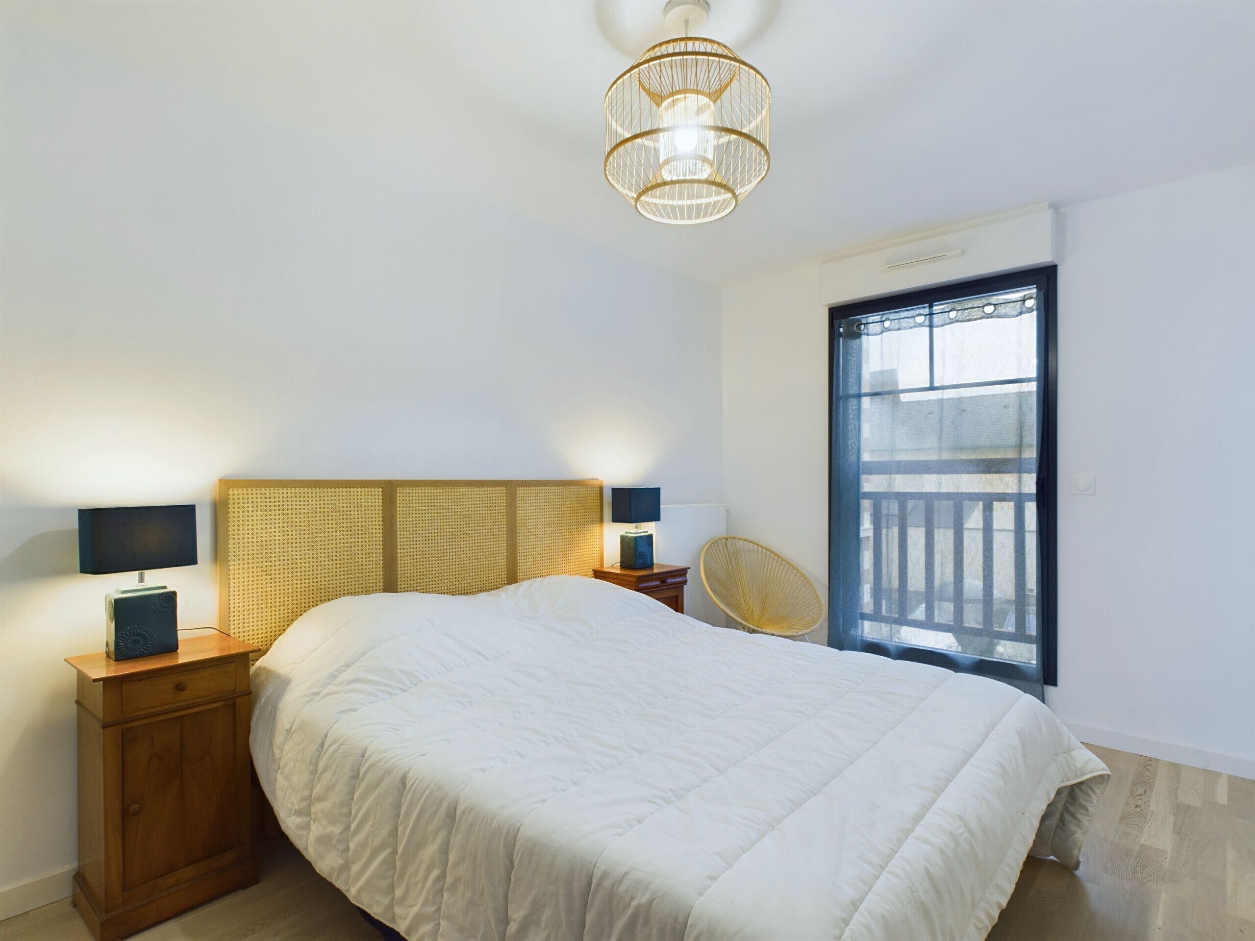 A bedroom with a bed, bedside lamps, a nightstand, a wicker chair, and a window with a view.