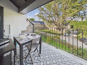 Un balcón con mesa y sillas, una parrilla y vistas a la calle y árboles.