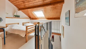 Quarto com teto inclinado, vigas de madeira, claraboia, cama, escrivaninha e uma pequena maqueta de barco à vela.