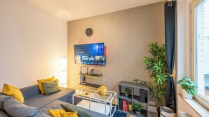 A modern living room with a grey sofa, a flat-screen TV, a bookshelf, and a potted plant. - Luminous Suite In Brussels - Elegant Design (Bruxelles)