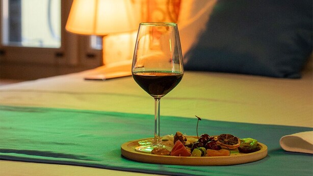 A glass of red wine on a plate with appetizers on a bed with a pillow and a lamp in the background. - Latibule Macaron Guest House (Ayvalik)