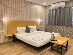 Habitación de hotel con cama de cabecera de madera, mesita de noche con teléfono, mesa con tetera y artículos de aseo, y ventana con cortinas.