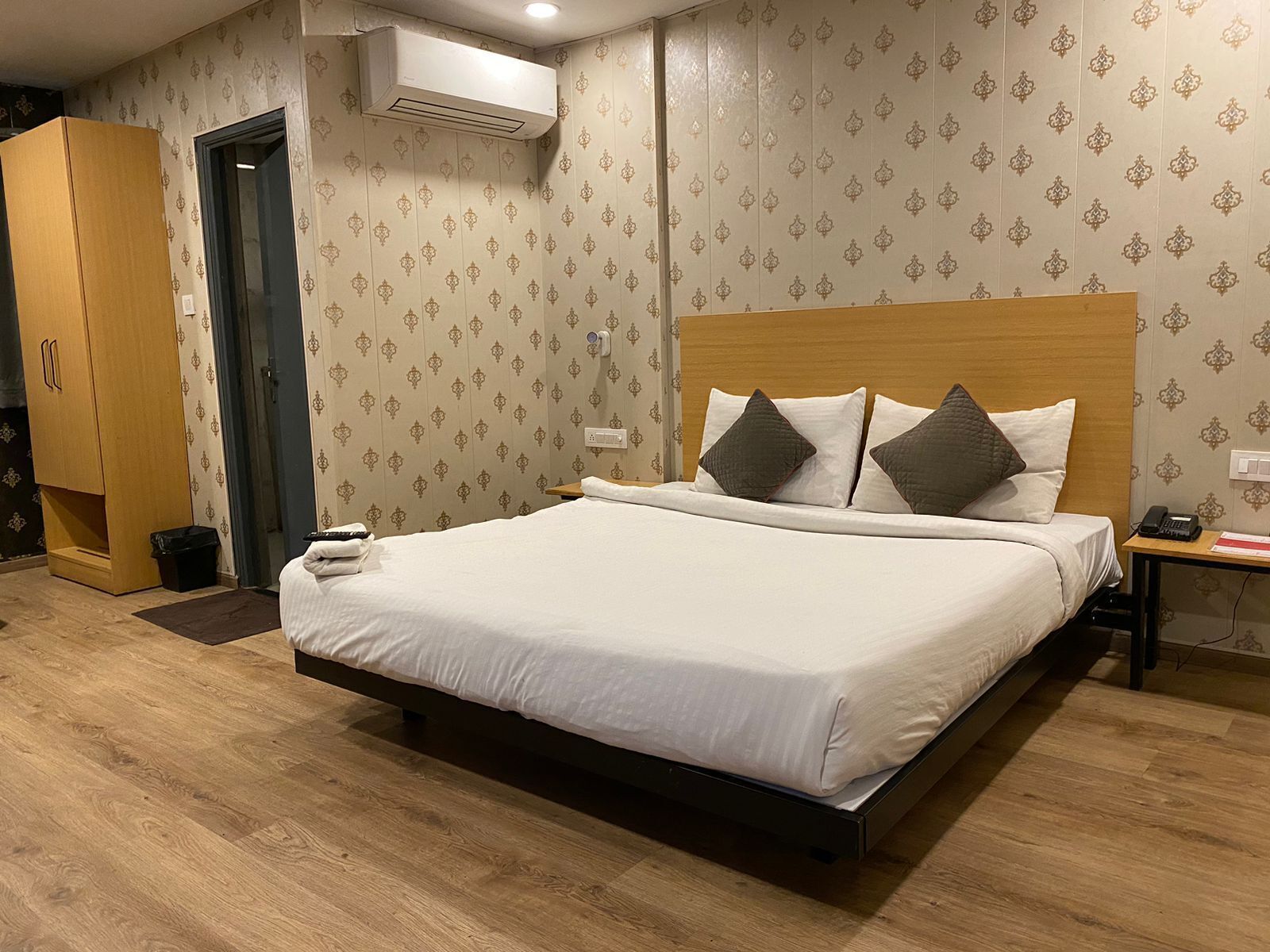 Een hotelkamer met een groot bed, een houten garderobe, een airconditioner en een nachtkastje met een telefoon en een rood boek.