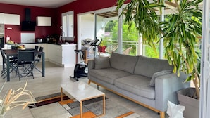 En moderne stue med en grå sofa, et hvidt sofabord og et spiseområde med bord og stole. Der er et stort vindue med udsigt til det fri.