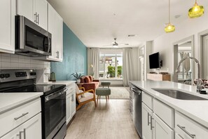 Una cocina moderna con armarios blancos, microondas integrado, estufa y lavabo. Hay una zona de estar con un sofá y una mesa pequeña.