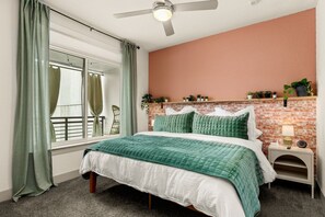 Un dormitorio con una pared de ladrillo decorativa, una cama con ropa de cama verde, una mesita de noche con una lámpara y una ventana con cortinas.