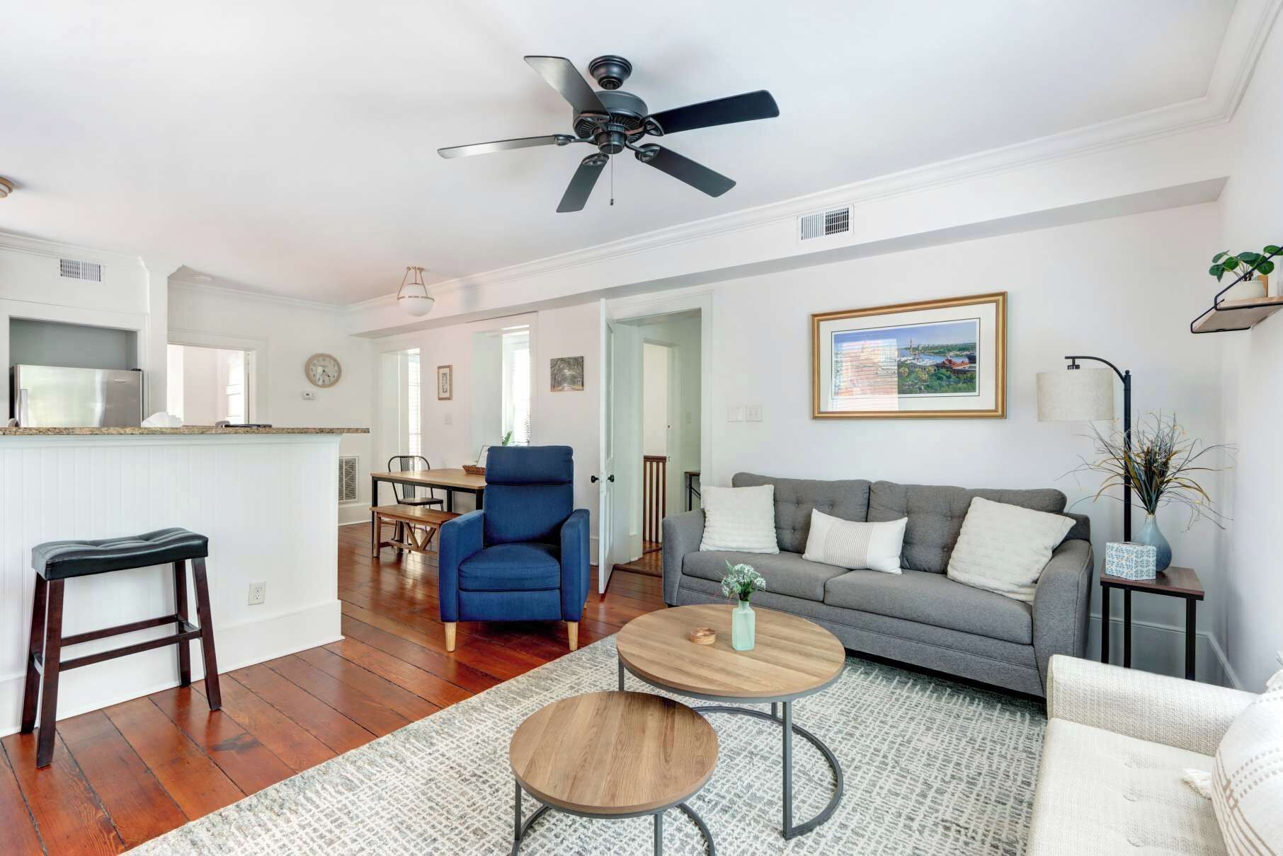 A spacious living area with a grey sofa, blue armchair, and round coffee table, a kitchen with white cabinets, and a ceiling fan.