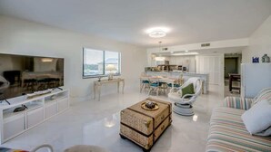 A spacious living area with a dining table, a sofa, a wicker ottoman, and a kitchen in the background.