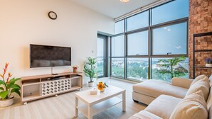 A modern living room with a large TV, a sofa, a coffee table, and a view of a cityscape through large windows.