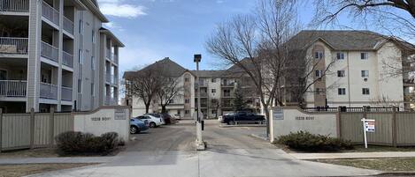 A gated residential area with multiple apartment buildings, a clear sky, and a parking lot with several cars.