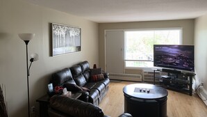 A living room with a black leather sofa, a coffee table, a flat-screen TV, and a painting on the wall. - Cozy condo, close to Northgate mall and Ring road Top Floor. (REGINA)