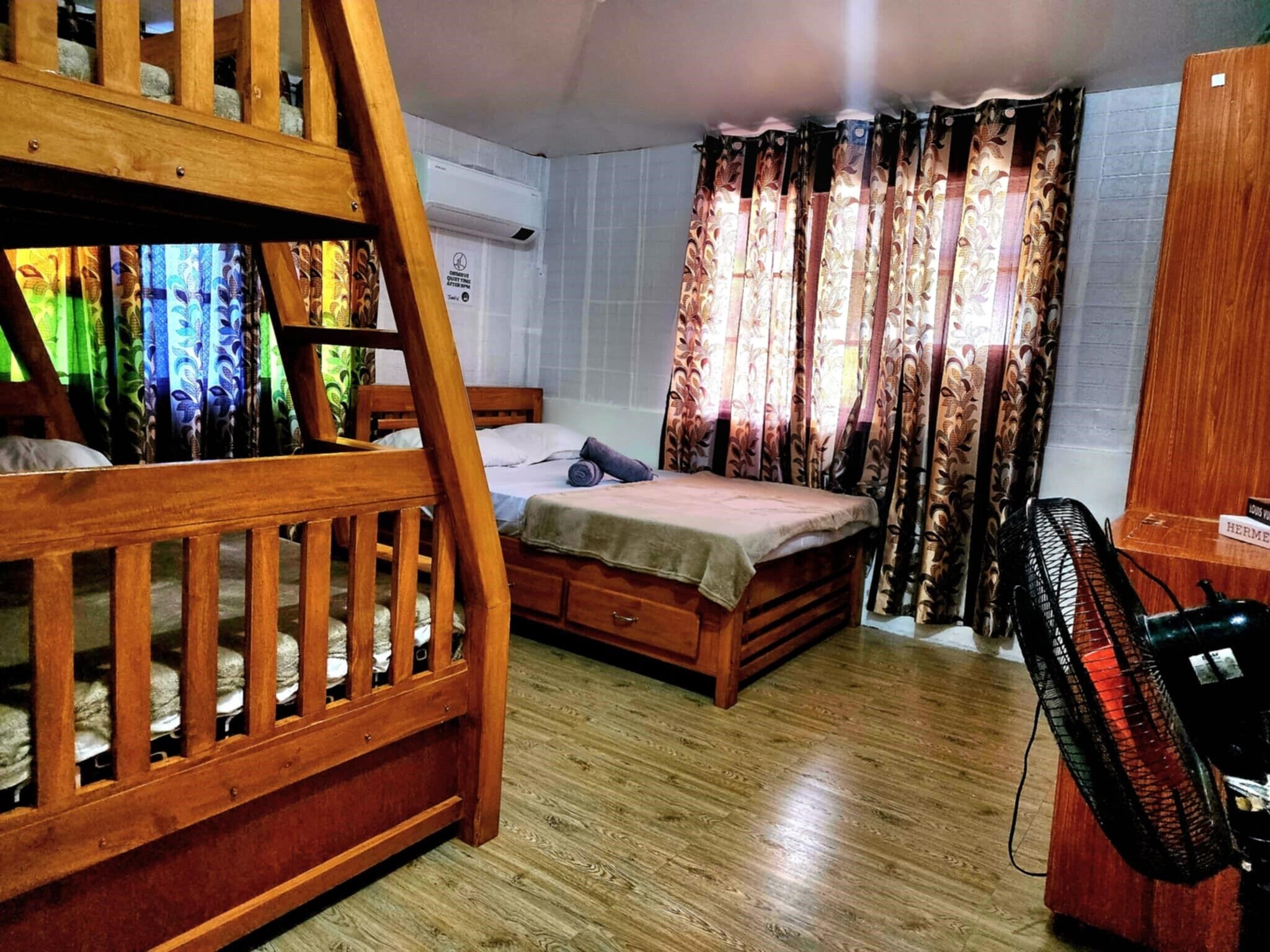 Una habitación con una cama litera, una cama individual, un armario de madera, un ventilador y cortinas estampadas.