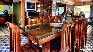 A dining area with a glass-top table surrounded by wooden chairs, a television, and a bar area with shelves of bottles. - 3 Bed House - Sleeps 20 - Pets - Pool - Garden (Aringay)