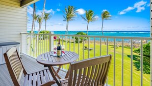 A balcony with a table and chairs, overlooking a beach with palm trees and the ocean. - Kapaa Shore A201 Oceanfront! (Kapaa)