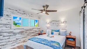 A bedroom with a wooden headboard, a ceiling fan, a bed with a blue and white patterned bedspread, a bedside table with a book, and a window with a view of greenery. - Kapaa Shore A201 Oceanfront! (Kapaa)