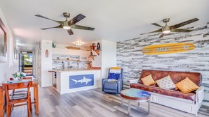 A modern living room with a brown leather sofa, a round coffee table, and a blue accent wall with a shark decal. A kitchen with white cabinets and a wooden island is visible in the background. - Kapaa Shore A201 Oceanfront! (Kapaa)