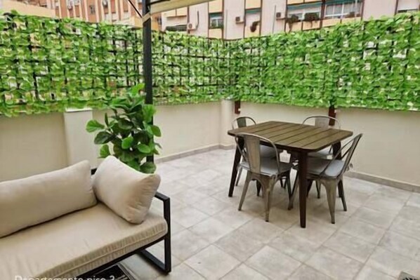 Un balcón con mesa y sillas de madera, un sofá color beige y una pared cubierta de follaje verde.