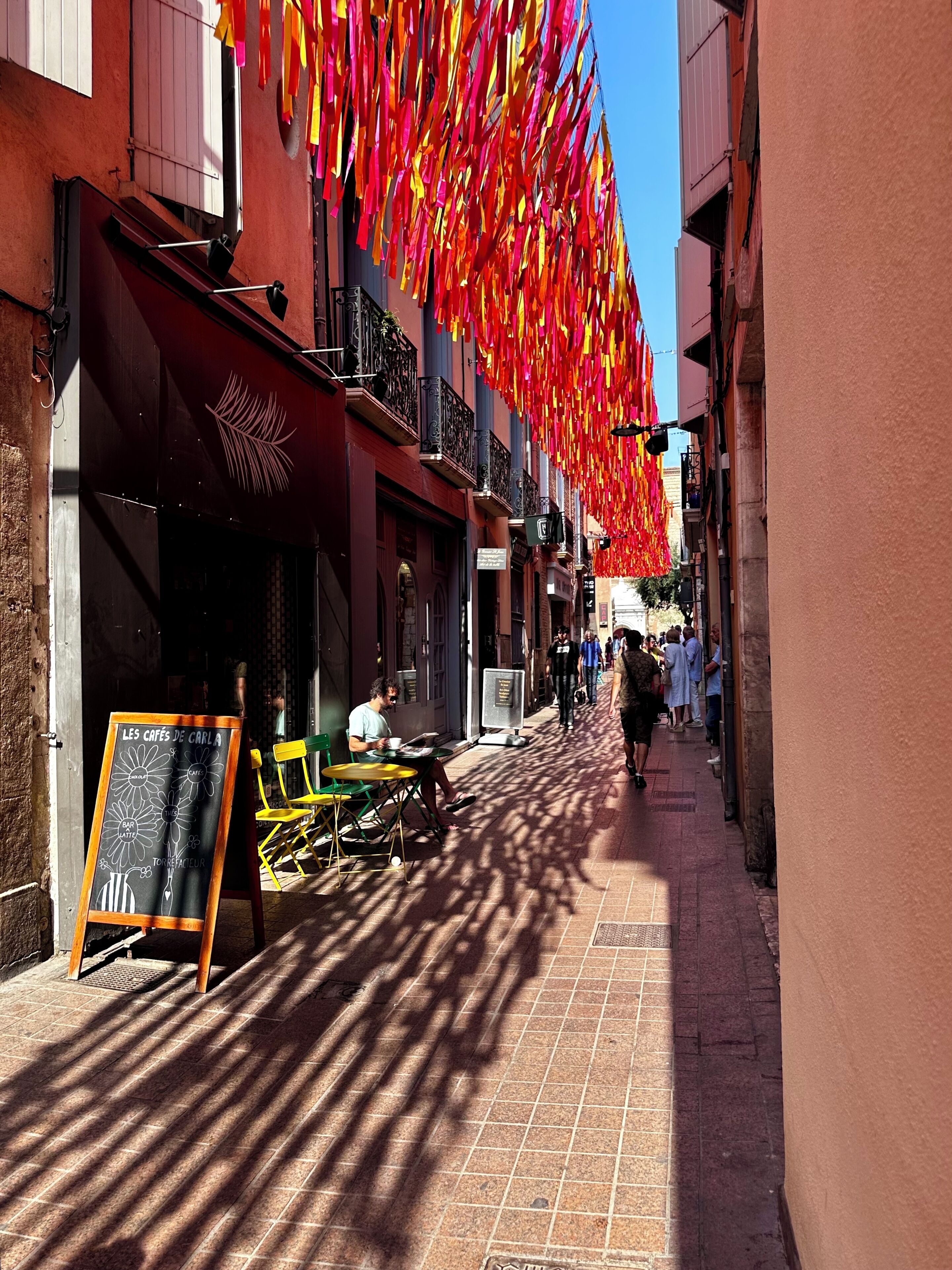 Une rue étroite, ornée de drapeaux rouges et jaunes suspendus au-dessus, un café avec des tables en terrasse et une enseigne publicitaire annonçant des cafés et des pâtisseries.