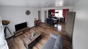 A modern living room with a sofa, coffee table, and TV. A kitchen is visible in the background with a dining area.