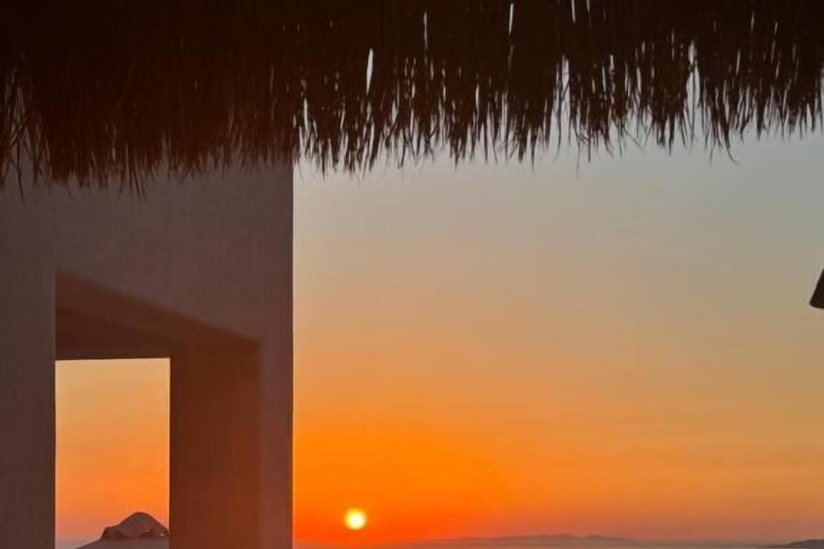 A sunset view from a balcony with a thatched roof, overlooking a calm sea and distant landforms.