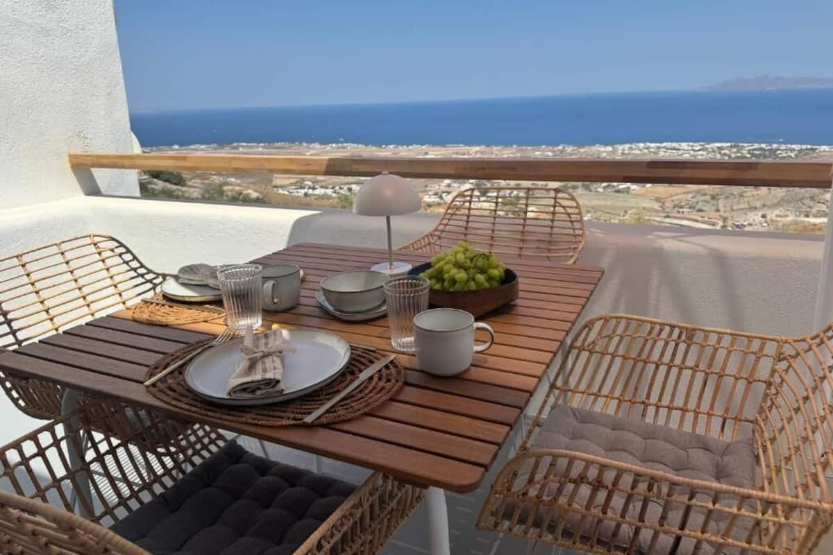 A balcony with a wooden table set for two, wicker chairs, and a view of the sea and cityscape.