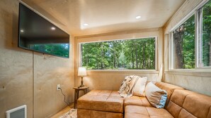 A cozy room with a large window, a flat-screen TV mounted on the wall, a cushioned bench, and a lamp on a side table. - Boho Bungalow-unique & cozy tiny home (Mentone)