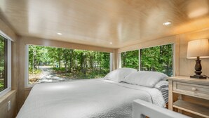 A bedroom with a large bed, a nightstand, and a window offering a view of a forest. - Boho Bungalow-unique & cozy tiny home (Mentone)