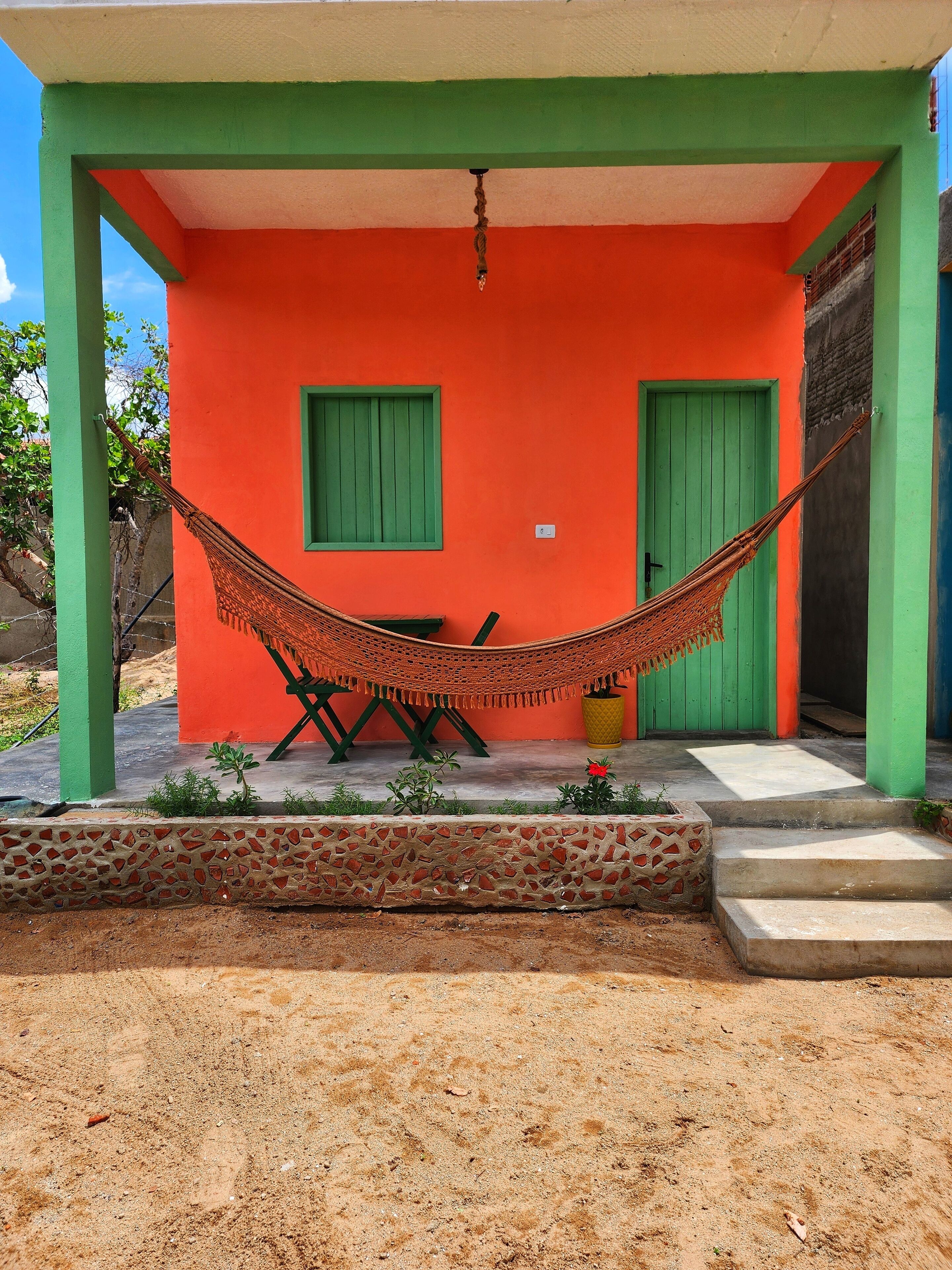 Un edificio colorido con una hamaca bajo un techo verde y naranja, una mesita y una puerta verde.