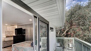 A balcony with a white railing, a bed with a floral bedspread, a TV on a dresser, and a view of trees and shrubs. - Great condo just a short walk to the beach (St. Simons Island)