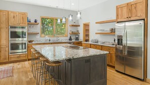 A modern kitchen with wooden cabinets, stainless steel appliances, a large island with stools, and pendant lighting. - Secluded 3BR home on 30 acres near Green River Gorge with hot tub & trails (Saluda)