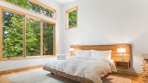 A bedroom with a large bed, two bedside tables with lamps, and a view of trees through large windows. - Secluded 3BR home on 30 acres near Green River Gorge with hot tub & trails (Saluda)