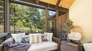Een houten terras met een bed, een hangstoel en uitzicht op een bos.