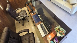 A modern office space with a wooden wall, a flat-screen TV, a refrigerator, a sofa, and a desk with a laptop and office supplies. - Hotel The Ceremony (Bhiwandi)