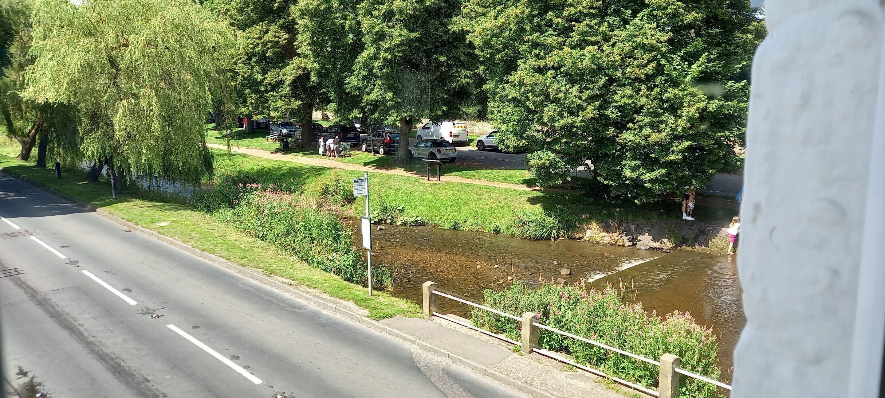 A road with a sidewalk and a small stream. There are trees and parked cars along the side of the road.