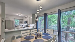 Una cocina moderna con una isla central, un comedor con mesa y sillas, y grandes ventanales con vistas a un bosque.