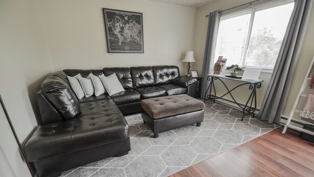 A living room with a leather sectional sofa, a matching ottoman, a hexagonal rug, a world map on the wall, a small table with a lamp, and a window with curtains.
