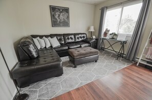 A living room with a leather sectional sofa, a matching ottoman, a hexagonal rug, a world map on the wall, a small table with a lamp, and a window with curtains.