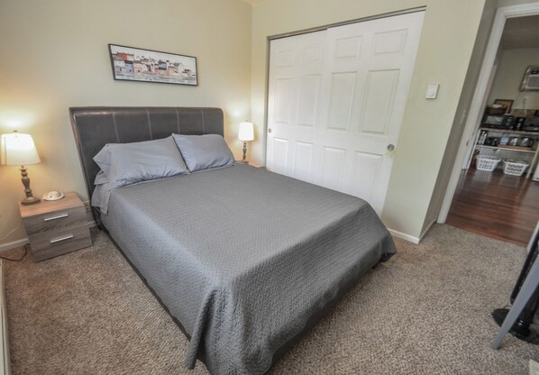A neatly made bed with a grey bedspread and pillows, a nightstand with a lamp, and a door to the right.