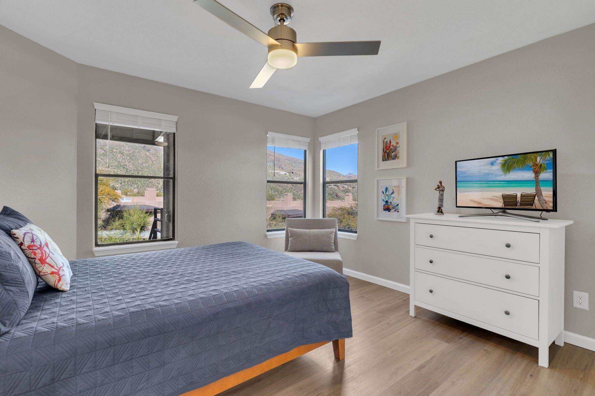 Una camera da letto con un letto, una ventola a soffitto, una TV, un comò e ampie finestre che offrono vista sull'esterno.