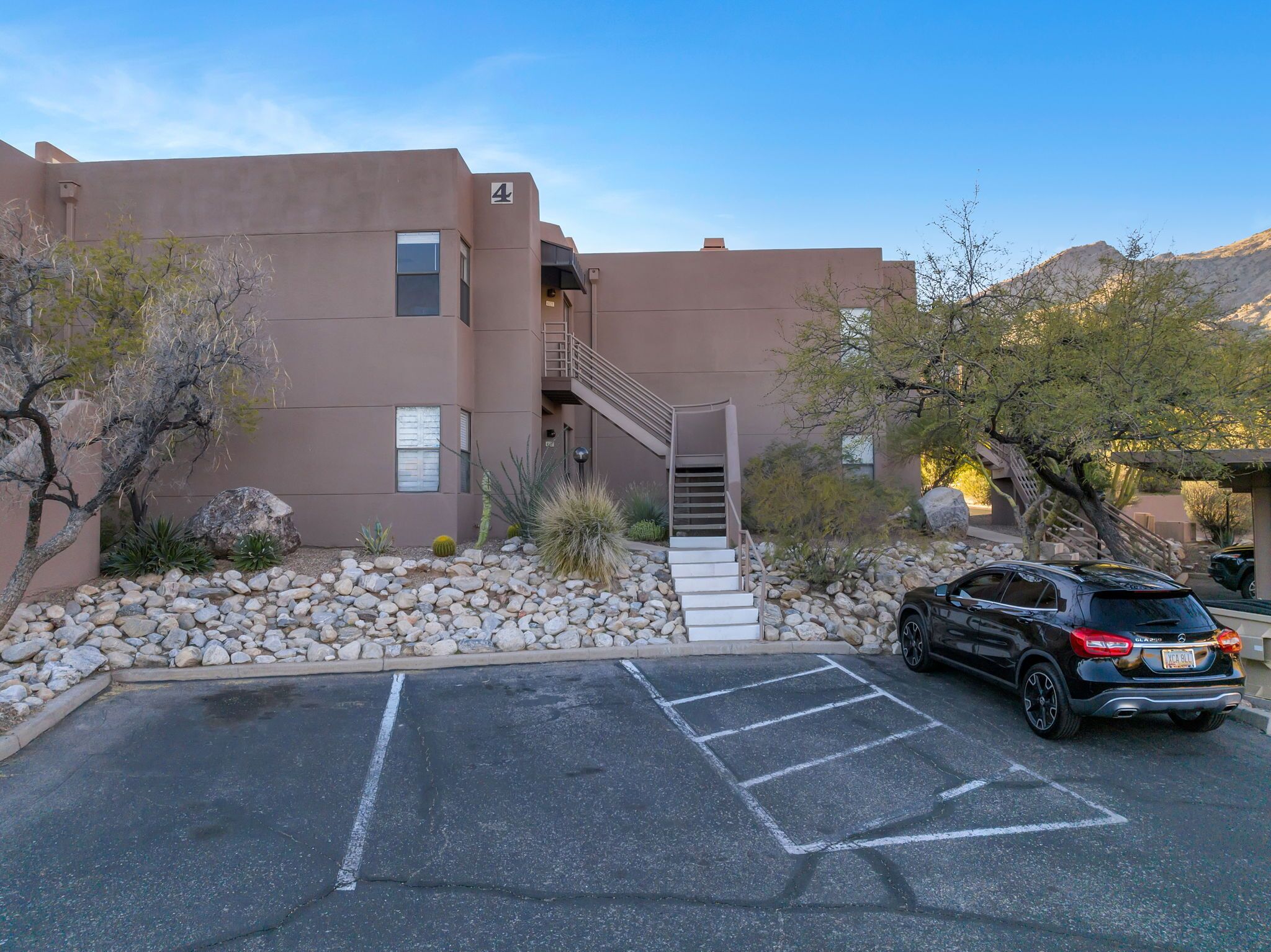 A building with a staircase leading to an upper level, a parking lot with a black SUV, and a desert landscape in the background.