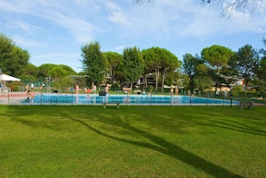 Ein Schwimmbecken, umgeben von Gras und Bäumen, mit einem Zaun und einigen Personen in der Nähe.