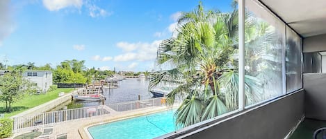 A balcony with a view of a swimming pool, palm trees, and a dock with boats.