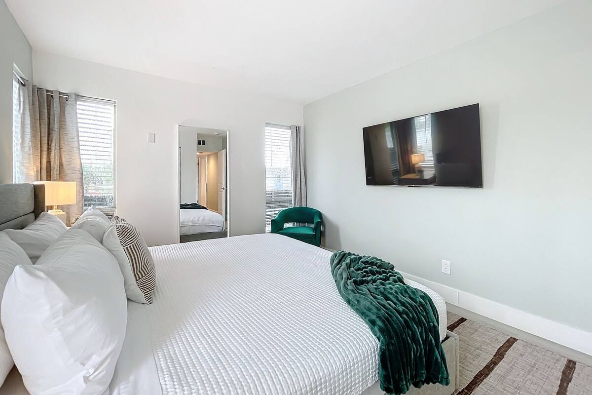 Habitación de hotel con una cama grande, un televisor de pantalla plana montado en la pared, una silla verde y una ventana con cortinas.